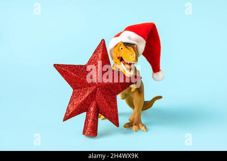 Dinosaurier Rex in rot Weihnachtsmann Hut hält Stern in den Pfoten auf blauem Hintergrund Silvester oder Heiligabend Art Weihnachtskarte Kreative Idee für Merry Stockfoto