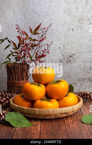 Süße Kaki im Bambuskorb. Persimmon Set Vorderansicht, Holztisch und vertikale Wand Hintergrund. Stockfoto