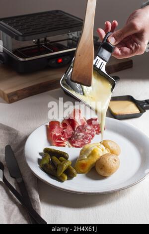 Hand hält ein Tablett mit geschmolzenem Raclette, das auf neue Kartoffeln auf einem Teller mit einer Seite von Charcuterie und Gurken gegossen wird. Stockfoto