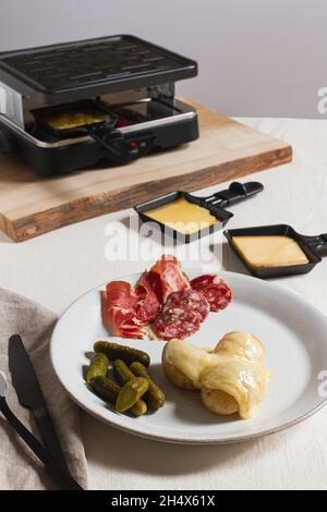 Geschmolzener Raclettekäse auf der Oberseite von neuen Kartoffeln auf einem Teller mit Charcuterie und Gurken. Stockfoto