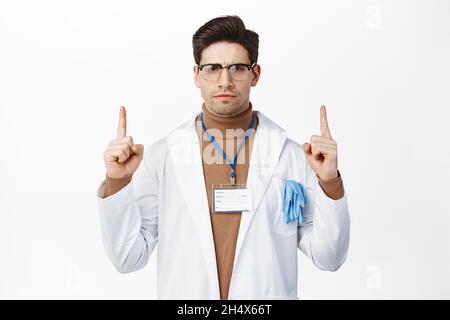 Wütender Arzt runzelte die Stirn und zeigte mit einem unzufrieden launischen Gesicht auf. Gesundheitshelfer in Robe mit Werbung auf weißem Hintergrund Stockfoto