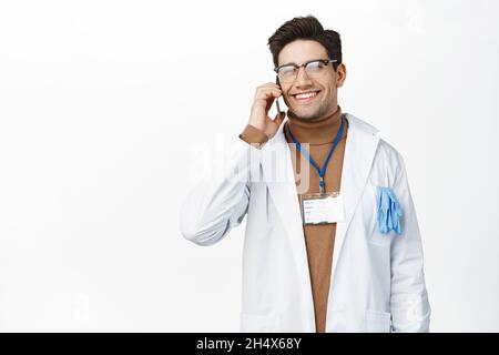 Lächelnder, männlicher Arzt nimmt Telefonanruf an, spricht über Handy, trägt Uniform-Gewand der Klinik und steht auf weißem Hintergrund Stockfoto