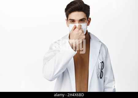 Der Arzt zieht eine medizinische Gesichtsmaske an und passt sie auf die Nase an, steht in der Uniform der Klinik, weiß isolierter Hintergrund Stockfoto