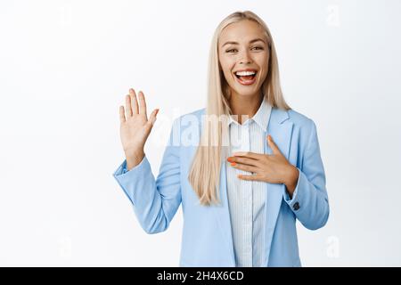 Junge Geschäftsfrau macht Versprechen. Die Verkäuferin hebt den Arm und legt die Hand auf das Herz, lächelt und sieht ehrlich aus und steht auf weißem Hintergrund Stockfoto