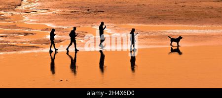 Ein Panoramabild einer Familie von Urlaubern, die am Fistral Beach in Cornwall spazieren gehen und von einem intensiven Sonnenuntergang umgeben sind. Stockfoto