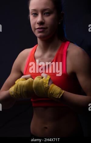 Junge und hübsche Boxerin, die ein rotes Trainingsplatten und gelbe Handhüllen trägt, ihre geballten Fäuste anstößt und mit einem leisen Lächeln auf die Kamera blickt. Anschluss Stockfoto