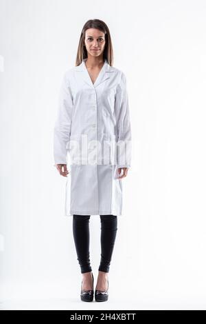 In voller Länge Porträt einer jungen Ärztin, die einen weißen Labormantel trägt und mit ihren Armen an ihrer Seite steht und leise auf die Kamera über Weiß blickt Stockfoto