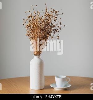 Tasse Kaffee und Vase mit flachsem trockenem Gras auf Holztisch. Weiße Wand auf dem Hintergrund. Stillleben nordische, skandinavische Wohnkultur. Gemütliches Wohnzimmer Stockfoto