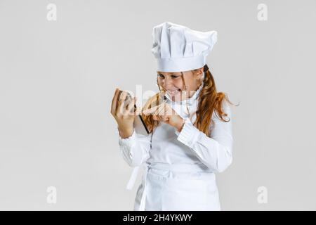 Lustige kleine niedliche Mädchen in weißen Koch Uniform und riesigen Koch Hut isoliert auf weißem Studio Hintergrund. Stockfoto