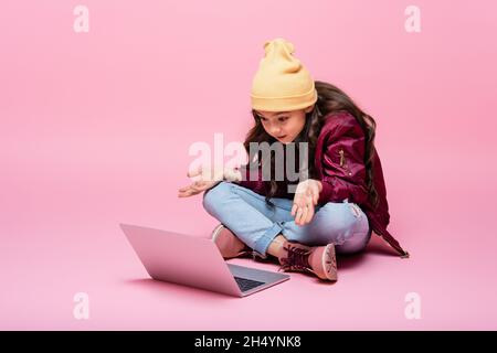 Stilvolle präteen Mädchen im Winter Outfit sitzen und Blick auf Laptop und zeigt mit Achselzucken Geste auf rosa Stockfoto