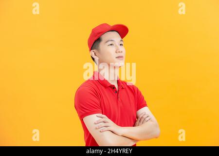 Lächelnder junger, hübscher asiatischer Entlieferungsmann in roter Uniform, der mit gekreuzten Armen steht Stockfoto