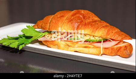 Croissant-Sandwich mit Schinken, Käse und Tomaten auf einem weißen Teller. Schwarzer Holztisch von hinten Close-Up. Fast-Food-Konzept, gesunde Ernährung. Stockfoto