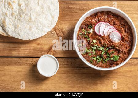 Traditionelles mexikanisches Gericht, Chili con Carne mit Hackfleisch, roten Bohnen, Kräutern und Tortilla in Papierform, serviert zum Mitnehmen auf dem Holztisch von oben Stockfoto