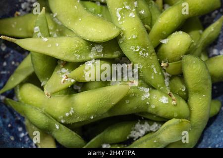 Biologischer gedämpfter japanischer Edamame, eine junge Sojabohne, die reich an Nährstoffen ist, serviert in einer blauen Schale und mit Meersalzflocken bestreut. Gemüse Backgr Stockfoto