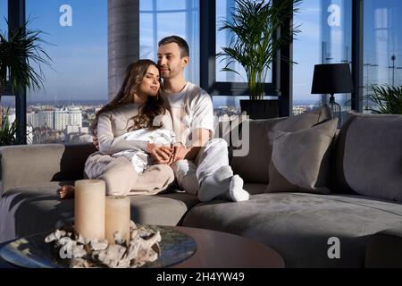 Attraktives junges Paar in legerer Kleidung, das sich zusammen auf der grauen Couch entspannt. Schöne Frau und Mann sitzen in Umarmungen in modernen Wohnung. Stockfoto