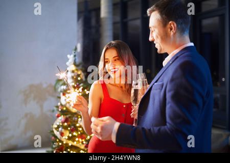 Glücklicher Mann und Frau halten funkelnde Strahlen und ein Glas Champagner, während sie sich am Abend gemeinsam entspannen. Junge Familie feiert Winterurlaub ay nach Hause. Stockfoto