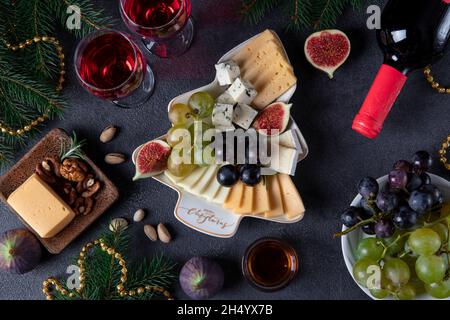 Verschiedene Käsesorten und Früchte, serviert auf einem Teller als Weihnachtsbaum, auf dunkelgrauem Hintergrund mit zwei Gläsern Wein. Draufsicht Stockfoto