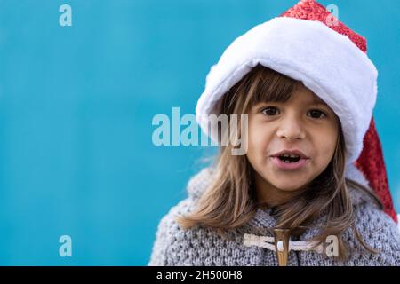 Kleines Mädchen trägt weihnachtsmann Hut Blick in die Kamera - Lustiges Kind spielt vor einer blauen Wand Hintergrund im Freien in der Weihnachtszeit - Frohe Weihnachten und h Stockfoto