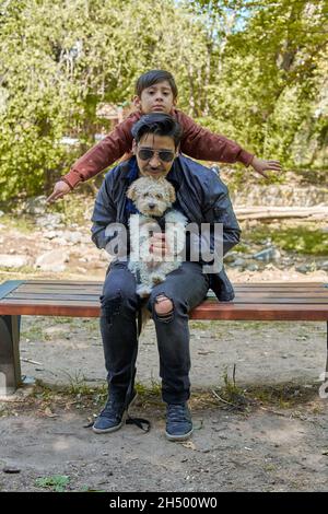 latino Vater und Sohn, und ein weißer Pudel Pudel Welpe sitzt auf einer Holzbank posiert für ein Foto auf dem anderen. Im Hintergrund ein out-of- Stockfoto