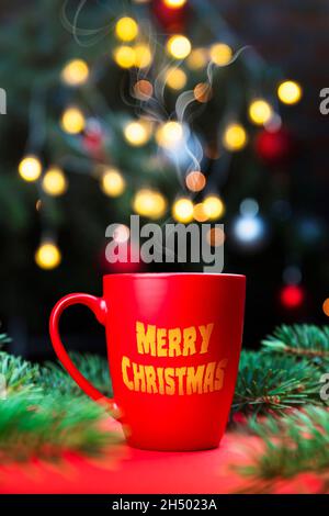 Köstlicher, frischer, festlicher Morgenkaffee in einer roten Tasse auf der roten Oberfläche mit Weihnachtsbaumlampen und Fichtenzweigen im Hintergrund Stockfoto