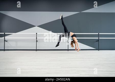 Seitenansicht einer Frau mit starker Passform, die Rad im Sprung übt und dabei in der Luft gespalten wird. Attraktives, flexibles Sportlertraining in der Tanzhalle mit Handläufen. Konzept der Choreografie, gesunder Lebensstil. Stockfoto