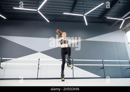 Junge hübsche kaukasische Mädchen in schwarzer Sportkleidung üben Tanzbewegungen, während kangoo springt und lächelt. Von unten sehen Sie die Sportlerin, die in der Halle Cardio-Übungen macht, Hightech-Innenausstattung. Stockfoto