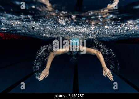 Professionelle Schwimmerin in Badekappe und Schutzbrille in Bewegung und Aktion während des Trainings am Pool, drinnen. Unterwasseraufnahmen Stockfoto