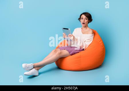 Full-Size-Profil Seitenfoto des jungen Mädchens erstaunt überrascht Sit Stuhl verwenden Tablette isoliert über blauen Hintergrund überrascht Stockfoto