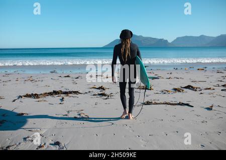 Rückansicht eines Mannes im Neoprenanzug, der mit Surfbrett am Strand in Richtung Kopierraum am blauen Himmel läuft Stockfoto