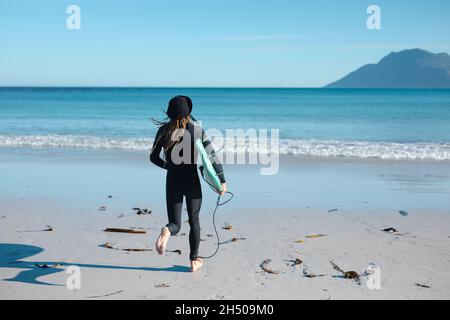 Rückansicht des männlichen Surfers, der an sonnigen Tagen mit Surfbrett an der Küste zum Meer läuft Stockfoto