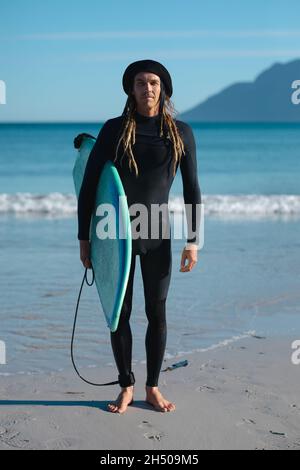 Porträt eines selbstbewussten männlichen Surfers in schwarzem Neoprenanzug, der an sonnigen Tagen am Strand Surfbrett trägt Stockfoto