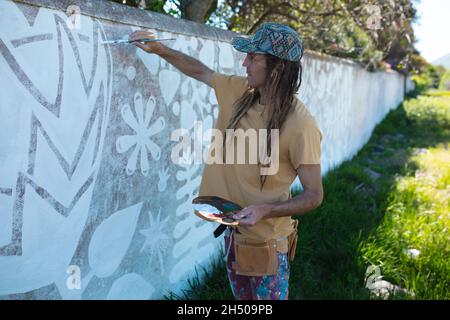 Männlicher Hipster-Künstler mit Palette, während er abstrakte Wandgemälde an die Wand malt Stockfoto