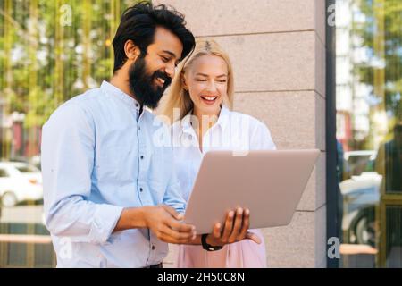 Seitenansicht eines fröhlichen bärtigen Geschäftsmanns und einer jungen attraktiven blonden Kollegin, die am Laptop arbeitet und im Freien etwas diskutiert. Stockfoto