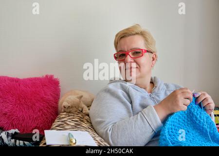 Porträt einer Frau, die auf einem Sofa sitzt und strickt und die Kamera anschaut. Ein süßes kurzhaariges Ingwer-Kätzchen nappt auf der Rückseite des Sofas Stockfoto