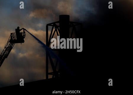 Hannover, Deutschland. November 2021. Im Licht der untergehenden Sonne löschen Feuerwehrleute das Feuer einer Industriehalle am Brinker Hafen von einer Drehleiter aus. Quelle: Moritz Frankenberg/dpa/Alamy Live News Stockfoto