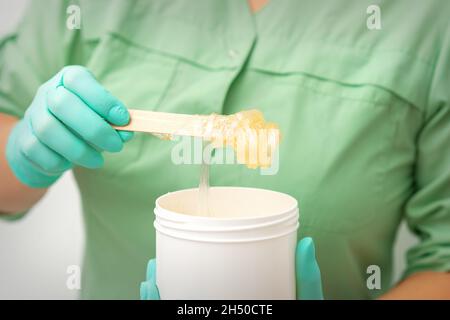 Flüssige gelbe Zuckerpaste zur Enthaarung auf einem Stock fließt in den Becher in den Händen einer Kosmetikerin Stockfoto