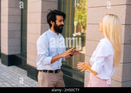 Seitenansicht einer attraktiven jungen blonden Geschäftsfrau und eines indischen bärtigen Geschäftsmannes, der im Freien durch ein Bürogebäude spricht. Stockfoto