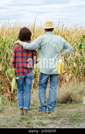 Kaukasische zweiköpfige Bauernfamilie im mittleren Alter mit Maiskolben in den Händen und Blick auf Maisfeld Stockfoto