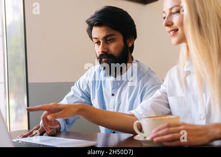 Nahaufnahme einer attraktiven Geschäftsfrau, die auf den Laptop-Bildschirm schaut und dem bärtig-hübschen männlichen Kollegen die Punkte erklärt Stockfoto