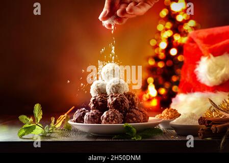 Von Hand geriebene Kokosraspel auf den Stapel von Kokosnuss- und Schokoladenkugeln auf den Weihnachtstisch und auf dunklem Hintergrund mit Tannenbaum, der mit Lig verziert ist Stockfoto