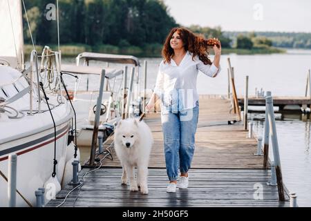 Eine glückliche Frau mit einem großen weißen Hund spaziert auf dem Pier in der Nähe der Yacht und des Meeres. Stockfoto