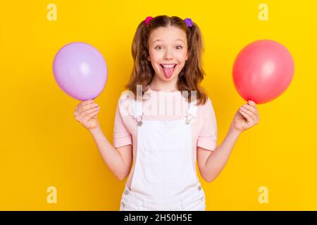 Foto-Porträt Schulmädchen mit Schwänzen zeigt Zunge halten Luftballons isoliert lebendige gelbe Farbe Hintergrund Stockfoto