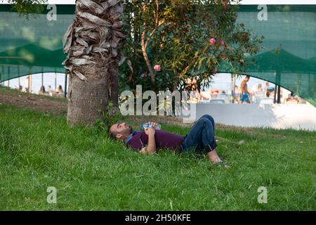 Kemer, Türkei - 08. 25. 2021: Ein Mann, der in der Sommersonne auf einem grünen Rasen einschlief. Ein Obdachloser schläft auf dem Gras im Park. Stockfoto