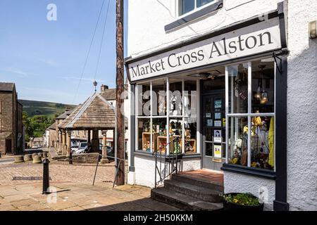 Ein Souvenirladen neben dem aus Stein erbauten Market Cross aus dem 18. Jahrhundert auf dem Platz der Upland Pennines-Stadt Alston, Cumbria, Großbritannien Stockfoto