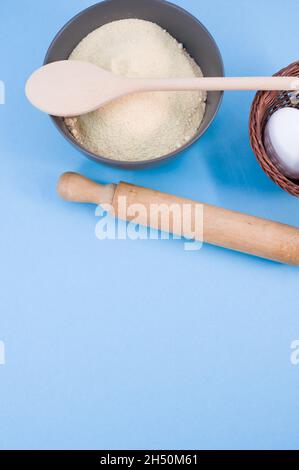 Dunkelgraue Schüssel mit Mehl Holzrollholz und weißen Eiern auf blauem Hintergrund Stockfoto