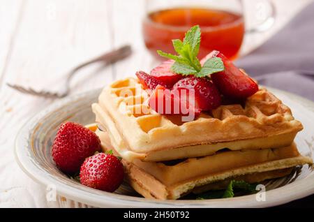 Blick aus der Nähe auf belgische Waffeln, serviert mit Erdbeeren und Minzblättern auf dem weißen Küchentisch aus Holz mit Sirup zur Seite Stockfoto