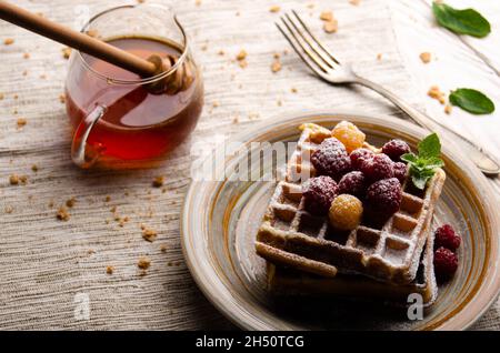 Belgische Waffeln serviert mit Himbeeren und Minzblättern, die mit Puderzucker bestäubt sind, auf dem weißen Küchentisch aus Holz mit Sirup zur Seite. Selektiver Fokus Stockfoto