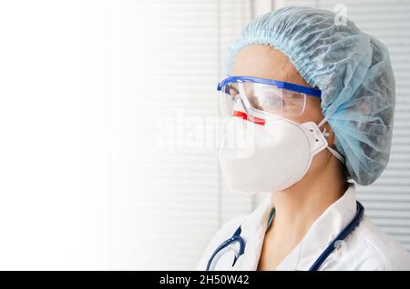 Porträt einer kaukasischen Ärztin in Maskenbrille und Hut mit Sternbild auf weißem Fensterhintergrund Stockfoto