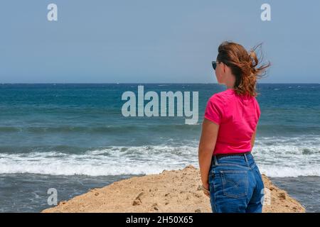 Wunderschöne, tausendjährige junge Frau, die eine blaue Jeans trägt, ein  leuchtendes rosafarbenes Oberteil und sonnendurchlässig auf die schaumigen  Wellen schaut, allein Reisende Frau Stockfotografie - Alamy