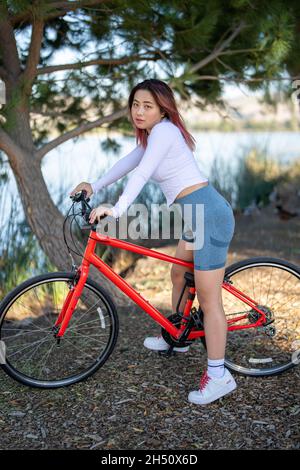 Junge asiatische Frau sitzt auf einem Orange Hybrid Fahrrad vor einem See Stockfoto